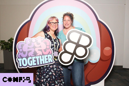 Two women smiling at the Figma Config 2024 design conference. They are standing in front of a colorful backdrop with a rainbow and holding playful Figma-themed props, including one that says 'TOGETHER.' Thea Eaton is on the right wearing a teal shirt, and Keira Dooley is on the left with glasses and a floral dress.